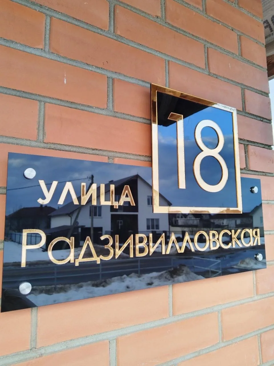 Черная глянцевая адресная табличка с объемными золотыми буквами «улица Радзивилловская» и номером 18 в золотой рамке, закрепленная на кирпичной стене