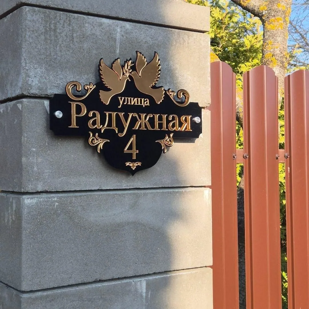 Фигурная черная адресная табличка с золотистыми буквами «улица Радужная», номером 4 и декоративными элементами в виде двух голубей с ветвью, закрепленная на бетонном столбе