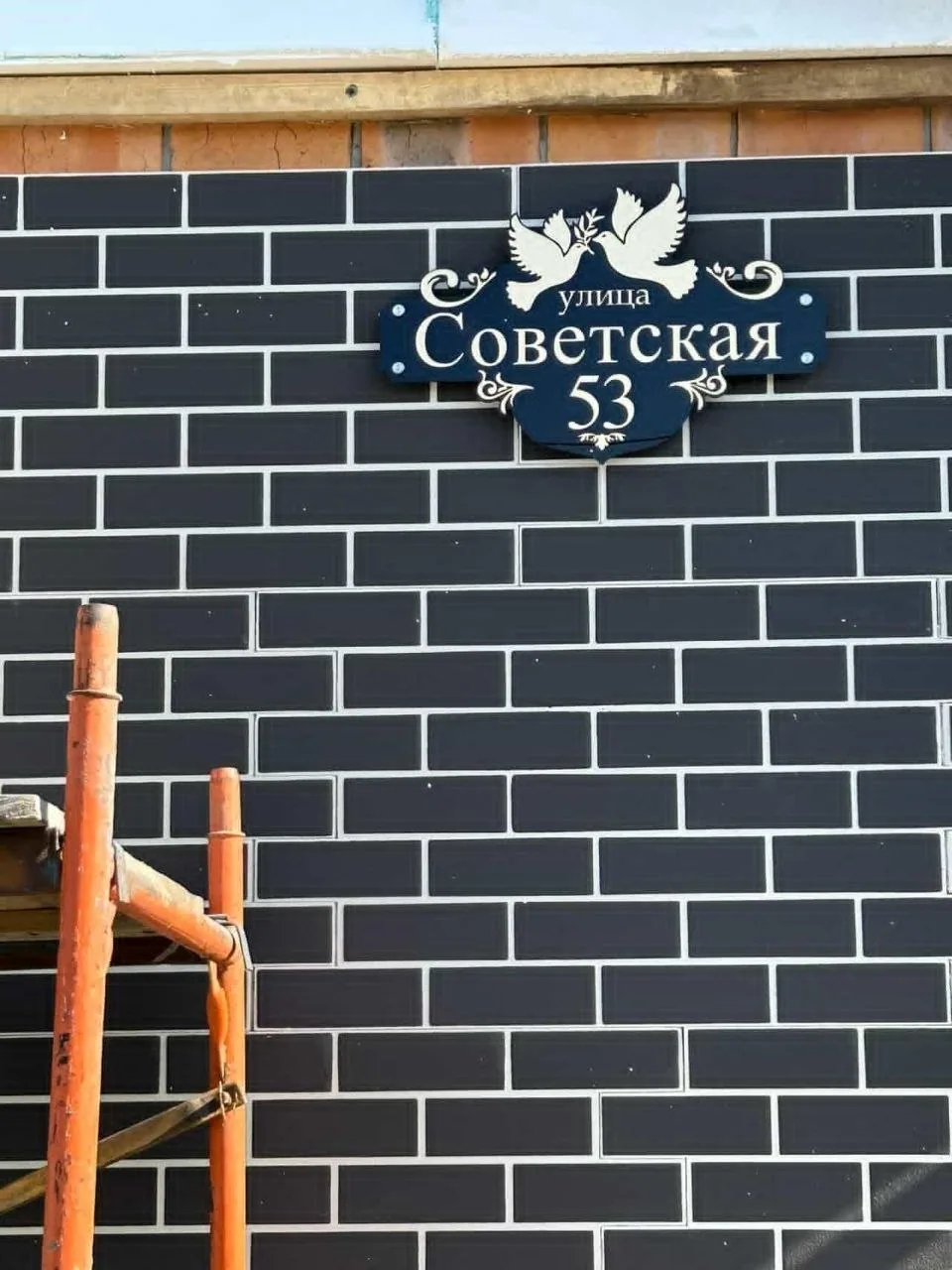 Темная фигурная адресная табличка с белыми надписями «улица Советская» и номером 53, украшенная объемным изображением двух голубей, на стене из темного кирпича с белой расшивкой
