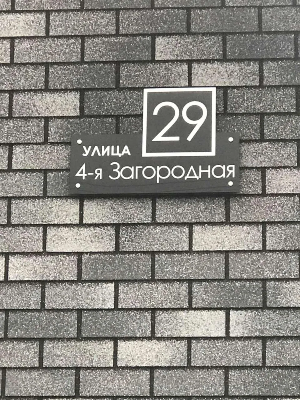 Черная двухсоставная адресная табличка с белыми надписями «улица 4-я Загородная» и крупным номером 29 в рамке, закрепленная на стене из серого декоративного кирпича