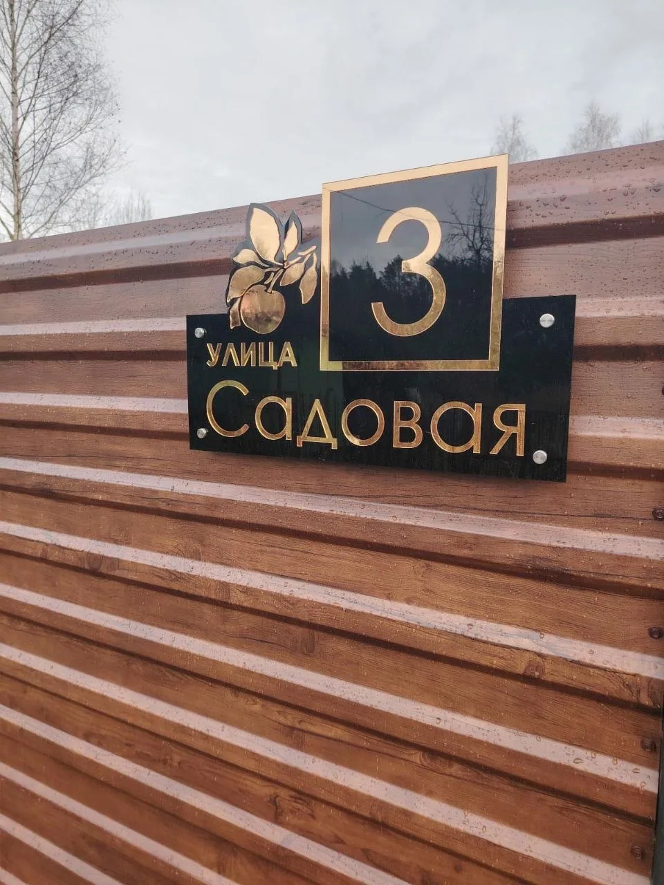 Черная глянцевая адресная табличка с золотыми надписями «улица Садовая», крупным номером 3 в рамке и объемным изображением яблока на ветке, установленная на коричневом заборе из профнастила